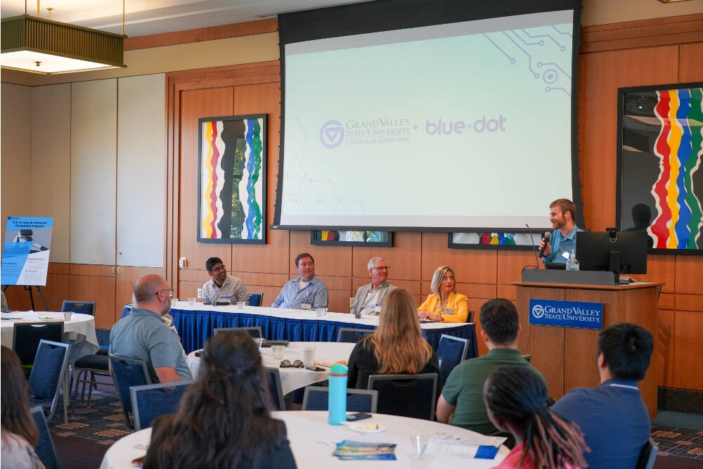 A speaker stands at a podium addressing an audience during a panel discussion at Grand Valley State University, with five panelists seated at a table on stage. The projector screen behind them displays the logos of the GVSU College of Computing and Blue D